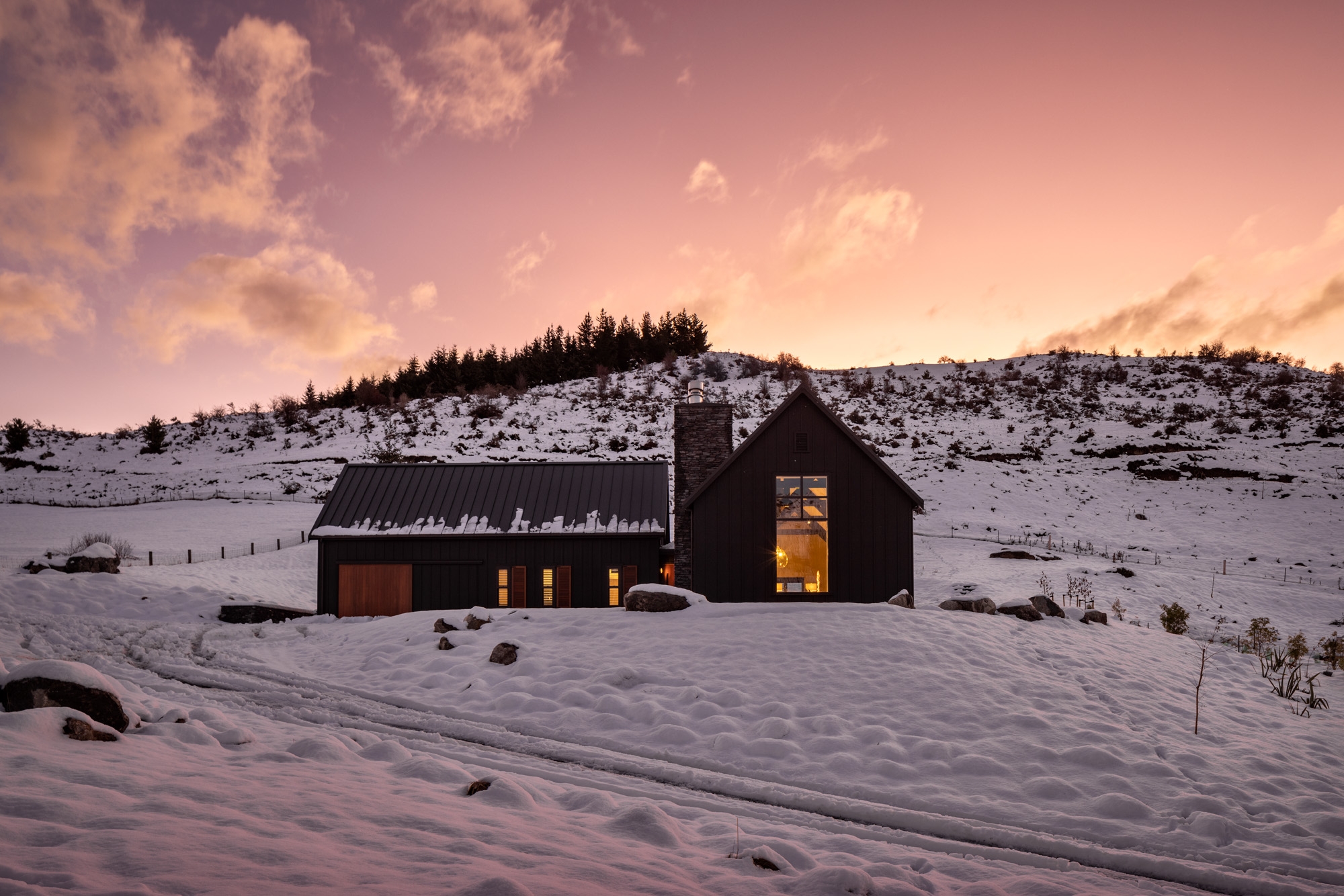 A modern black barn house sits in a snowy landscape against a backdrop of hills during sunset, with warm light glowing from its windows.