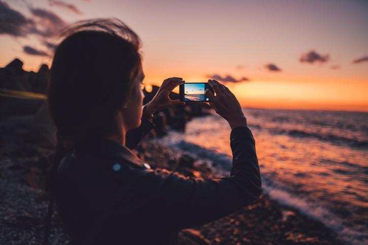 Woman at New Zealand beach photographing the sunset on an iPhone 14