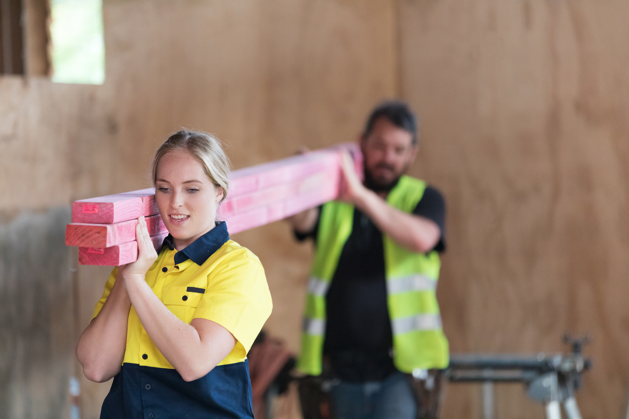 Female labourer carrying plywood planks over her shoulder along with male colleague