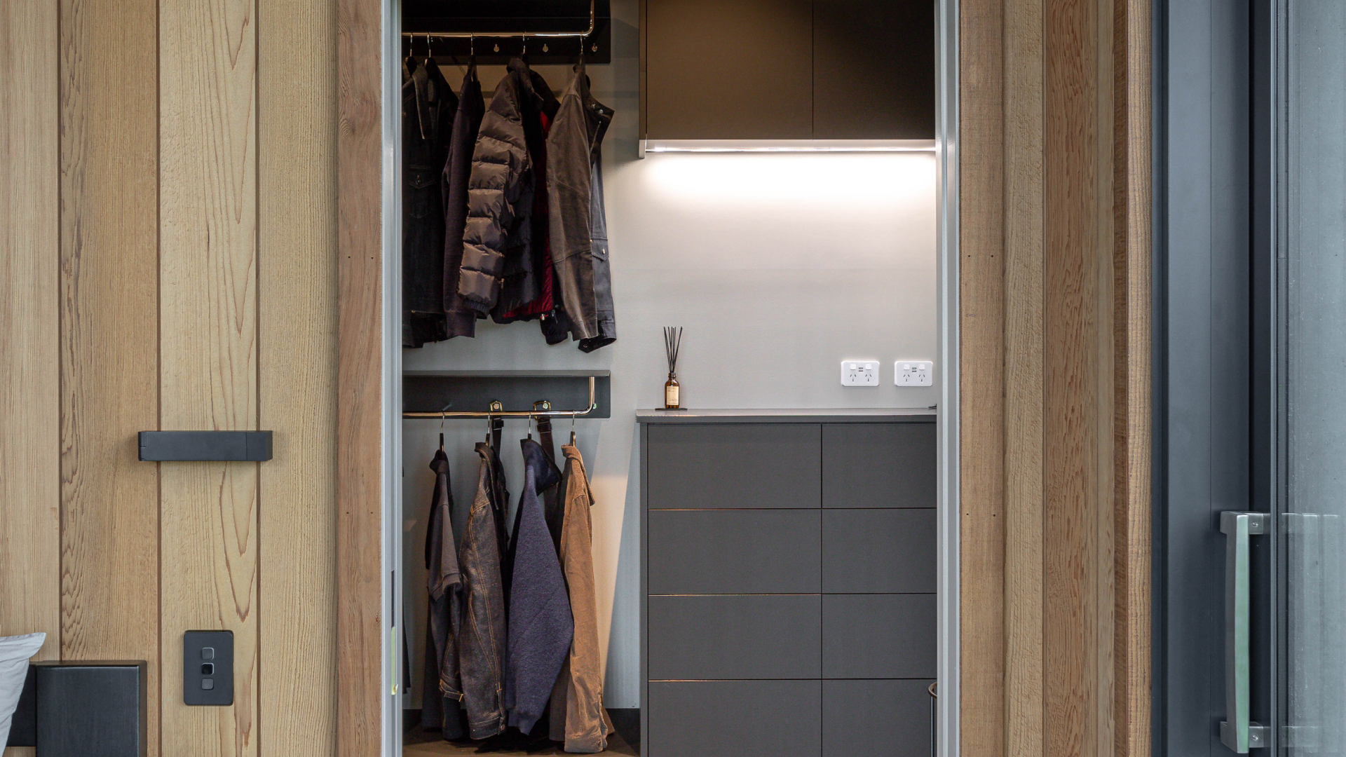 A walk-in wardrobe with built-in shelving featuring dark grey cabinets and a hanging area for clothes. The closet is viewed from a bedroom with wood-paneled walls and terrazzo flooring, creating a sleek and modern aesthetic.