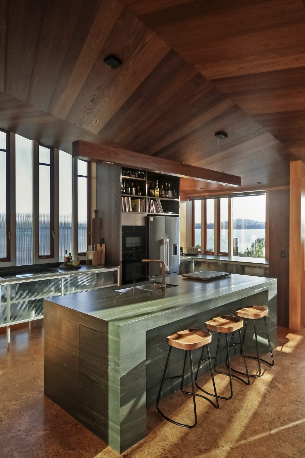Green kitchen island with three stools. High wooden roof and views of Auckland harbour.