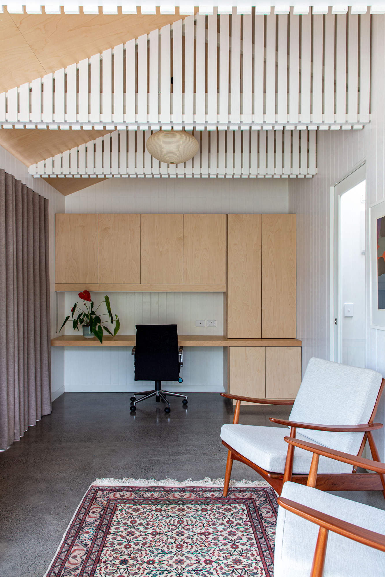A minimalist office space with concrete floors and wooden cupboards. A red rug in the centre of the floor with two chairs for relaxing around it. On the back wall is a inbuilt desk space which has been built in a light wood. 