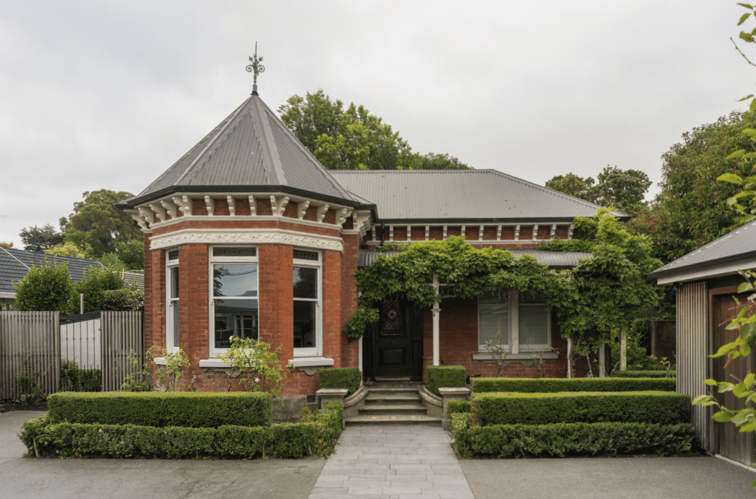 The image depicts a charming single-story residential building with a red brick facade. It features a bay window, a pointed roof adorned with decorative trim, and a black door flanked by columns. Landscaping surrounds the entrance path, and the sky appears overcast. This architectural style might be relevant for real estate listings or discussions about home design.