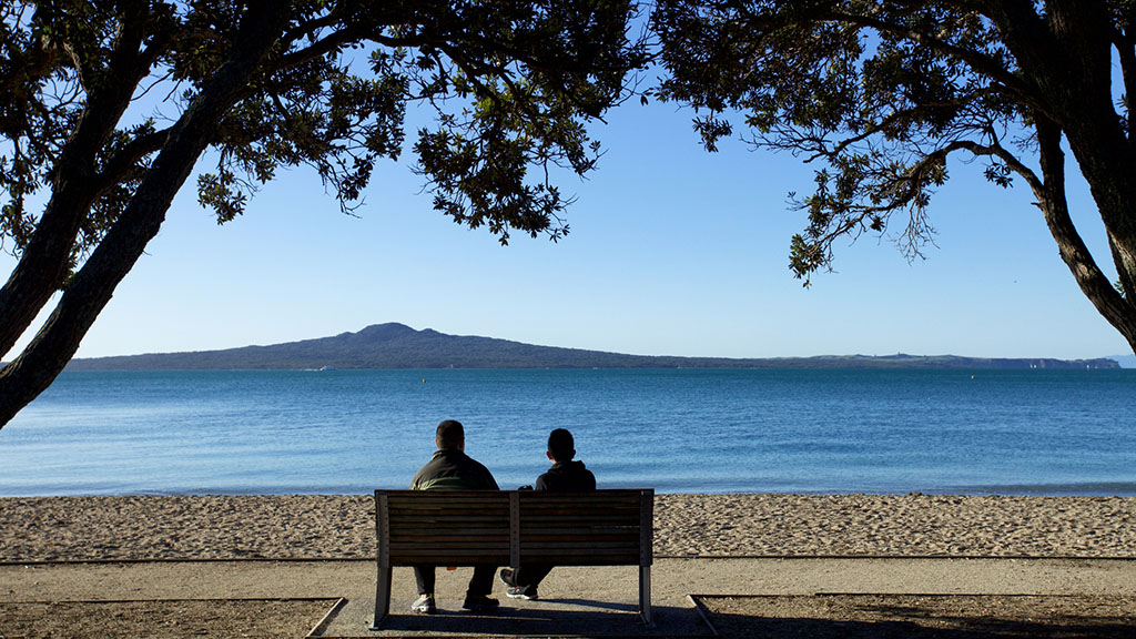 Two people on bench seat at Mission bay, looking out to Rangitoto Island