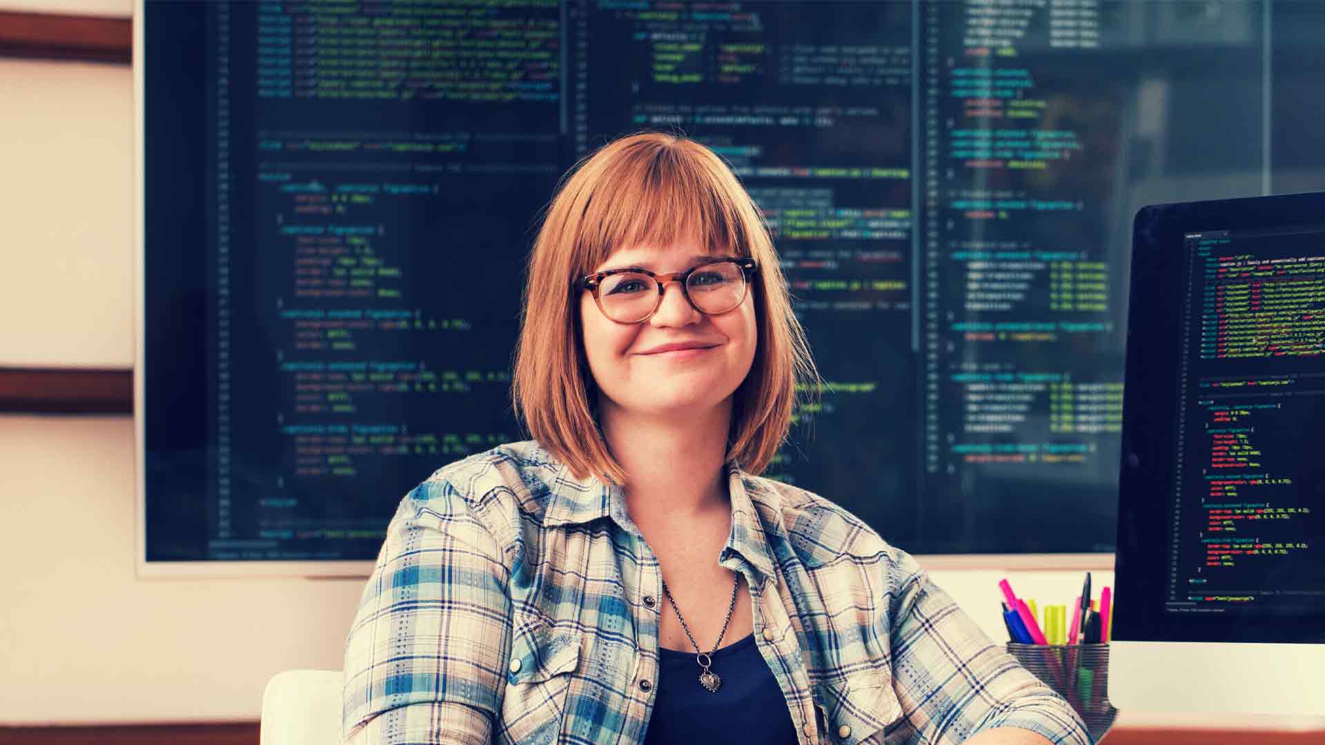 Smiling IT employee with screens with code behind her.