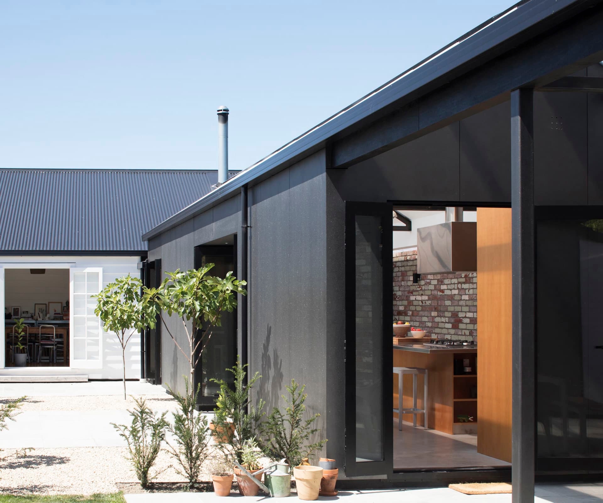 The image depicts a modern single-story house with a flat roof design. The exterior walls are painted in a dark color, possibly black or charcoal. There’s an open sliding door leading into the interior, revealing a glimpse of the kitchen area with wooden cabinets and a brick backsplash. Outside, several potted plants adorn the entrance, and a small tree is planted in the ground. The clear blue sky suggests it might be a sunny day. This image could be relevant for architectural design interests or real estate showcases due to its focus on modern home design and indoor-outdoor living spaces.