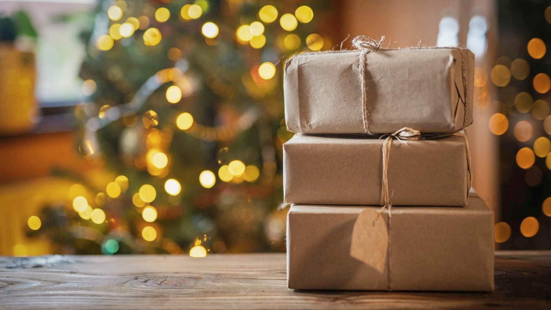 Stack of three Christmas present stacked on top of each other and wrapped in brown paper on top of a wooden table with a Christmas tree in the background.