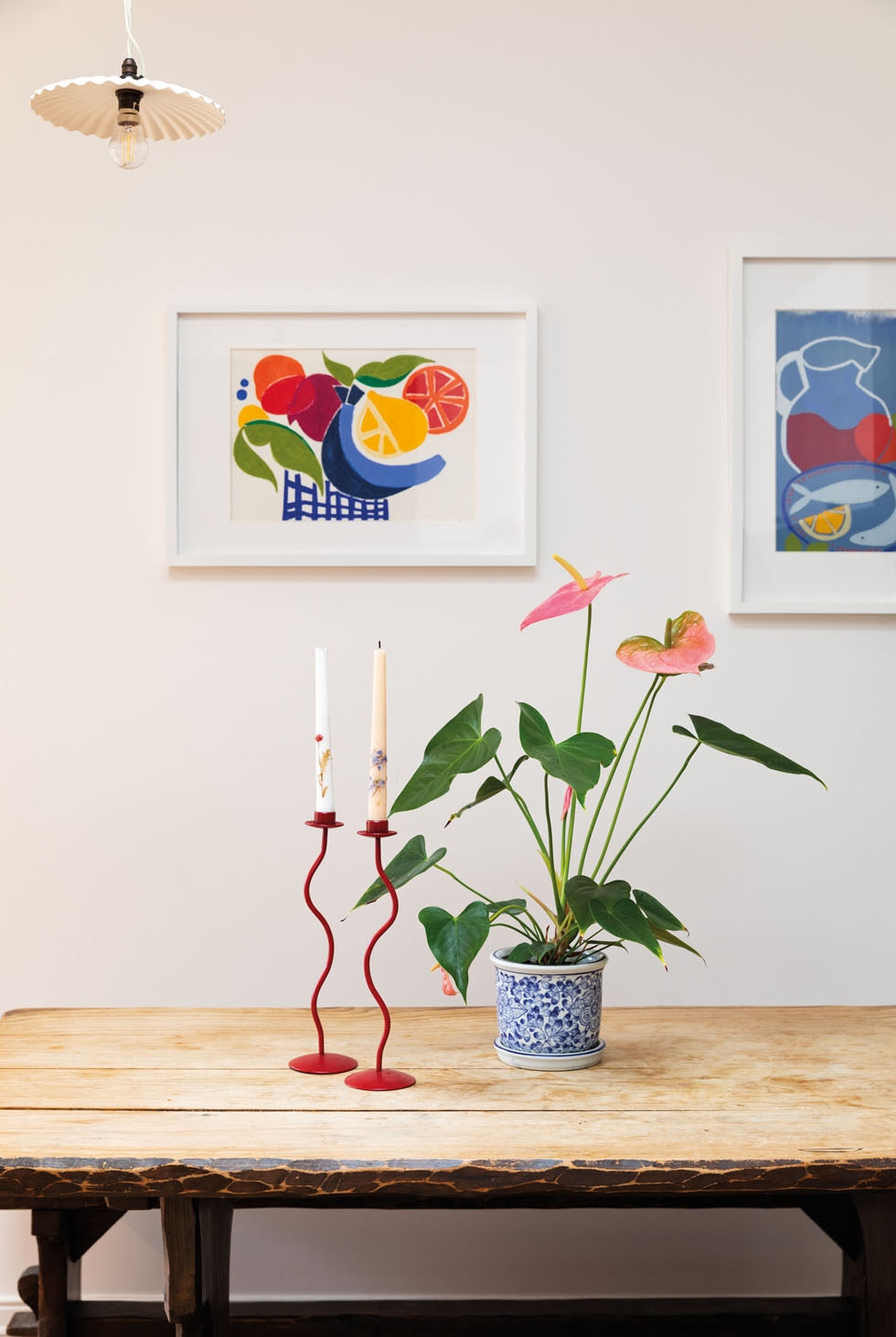 A set of candlesticks and a potted plant sit atop a wooden table, with art on the wall behind