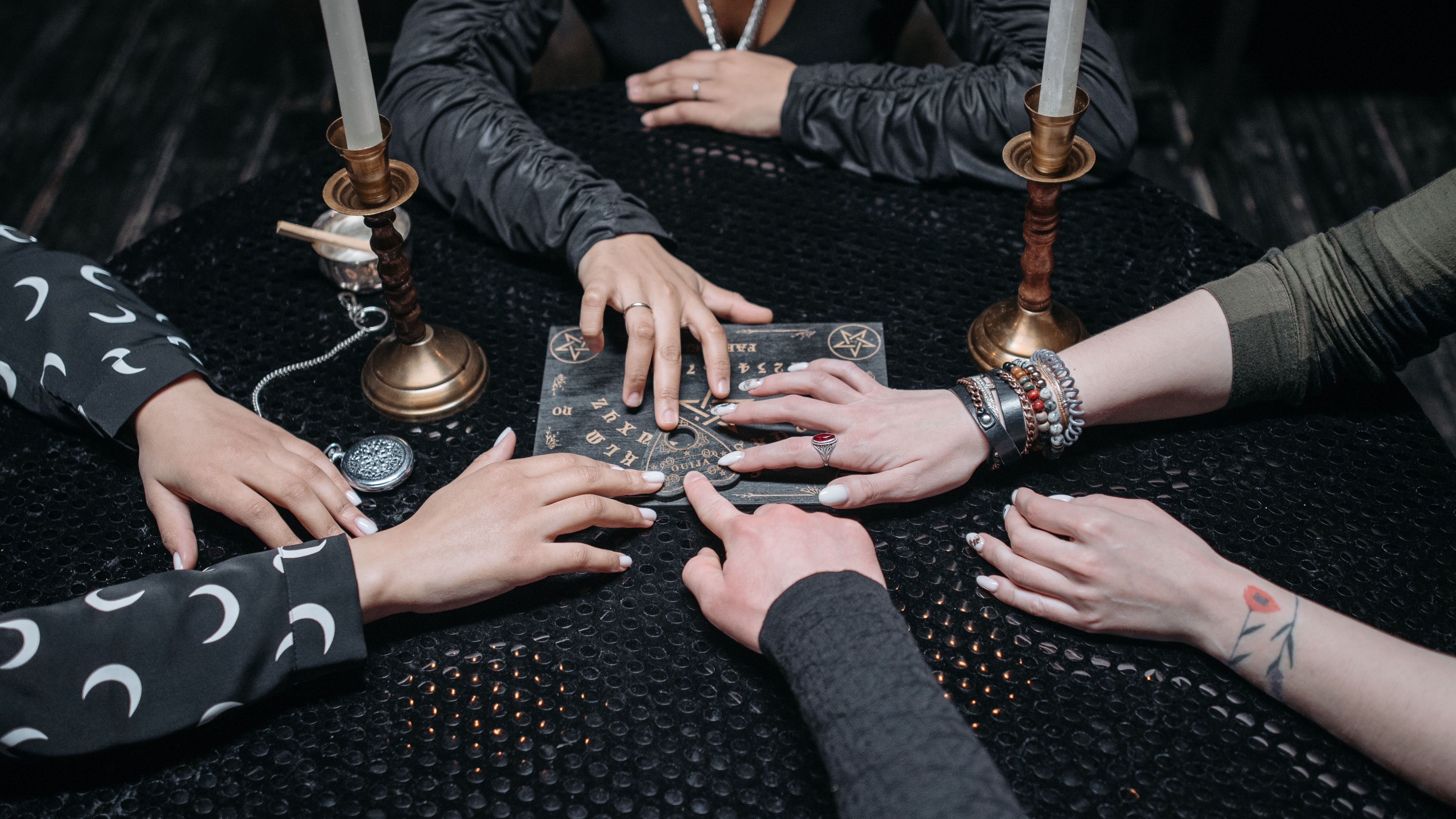 Four friends sitting around a table using a Ouji board. The table has a black table cloth and tall white candles on it.
