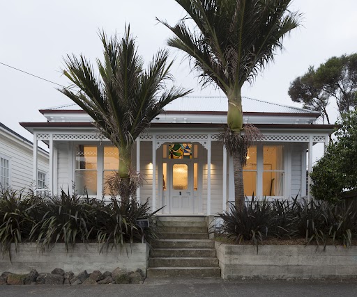 Image shows front of a white New Zealand villa with lights on in the house and two trees outside