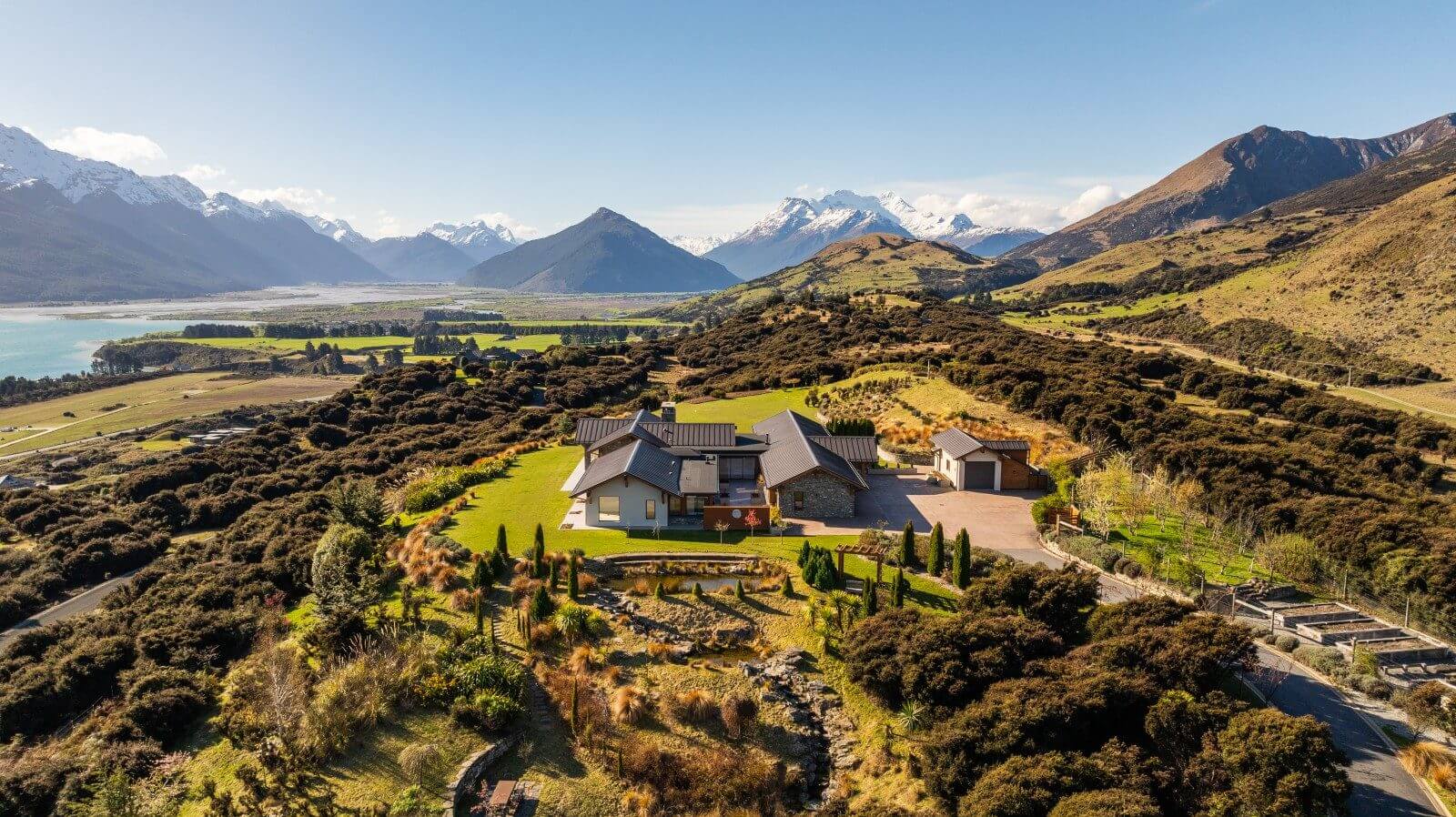 Aerial view of contemporary home in mountain valley with turquoise lake, snow-capped peaks, and native scrubland