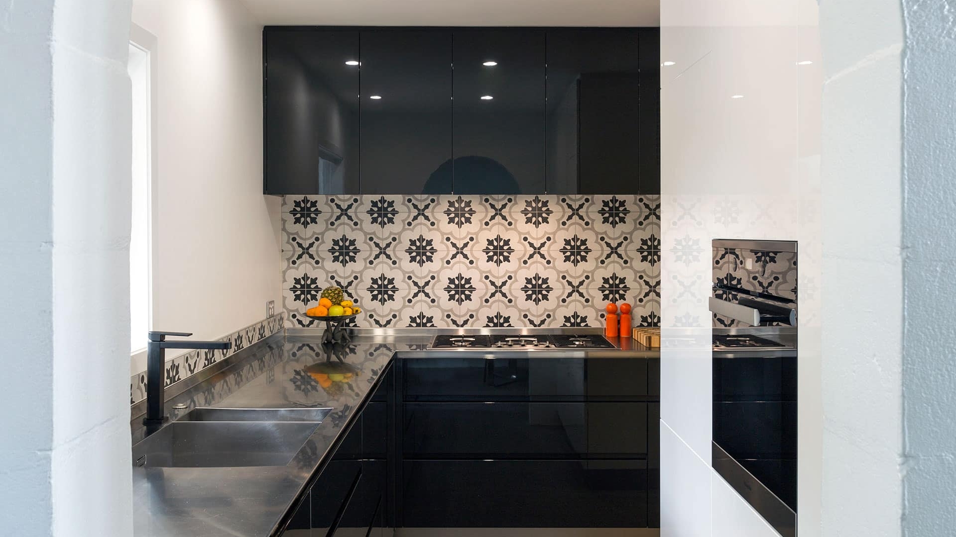 A modern kitchen with a black stovetop integrated into a dark countertop, white and patterned tile backsplash, and a black range hood against a black wall with cabinets.