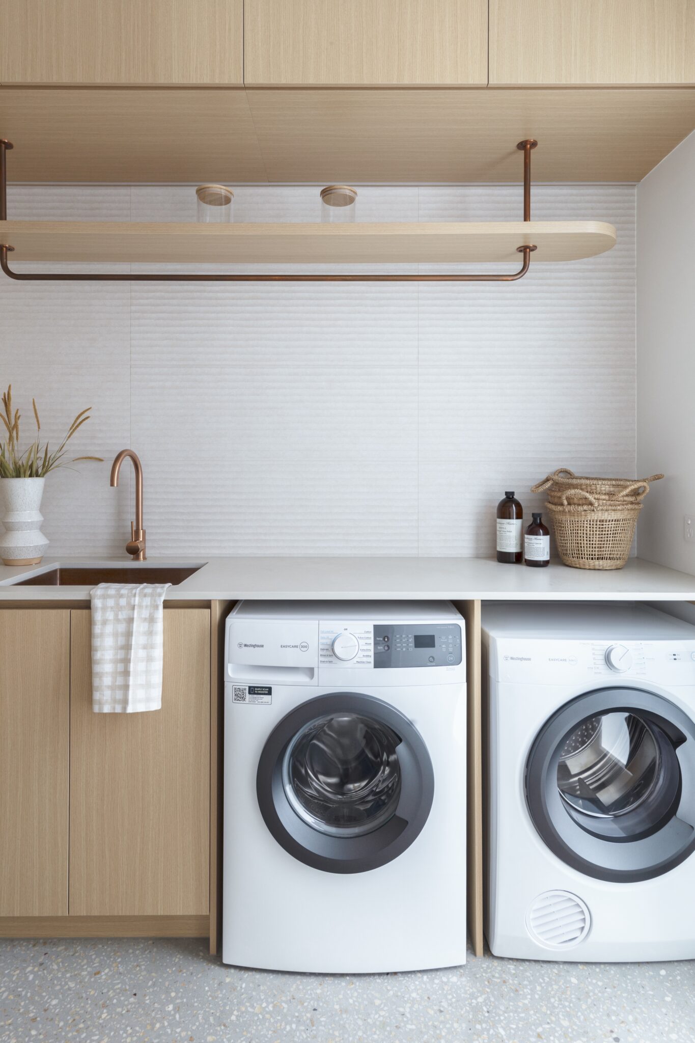 A sleek laundry room with a wooden and white design, featuring front-loading appliances and a minimalist clothing hanger for added functionality.