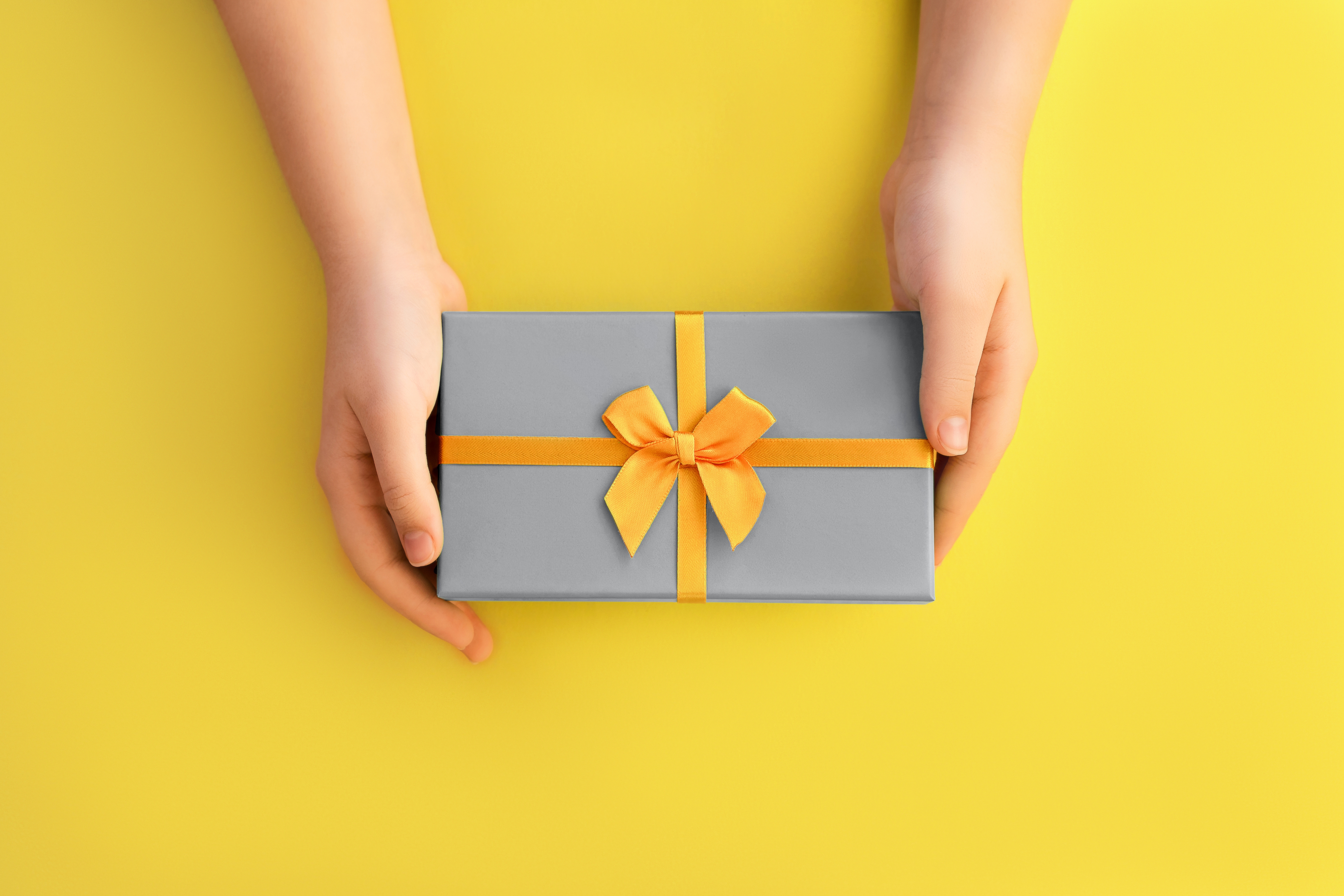 Top gift ideas for boyfriend. Two hands holding a grey gift box with yellow ribbon, on a yellow background.