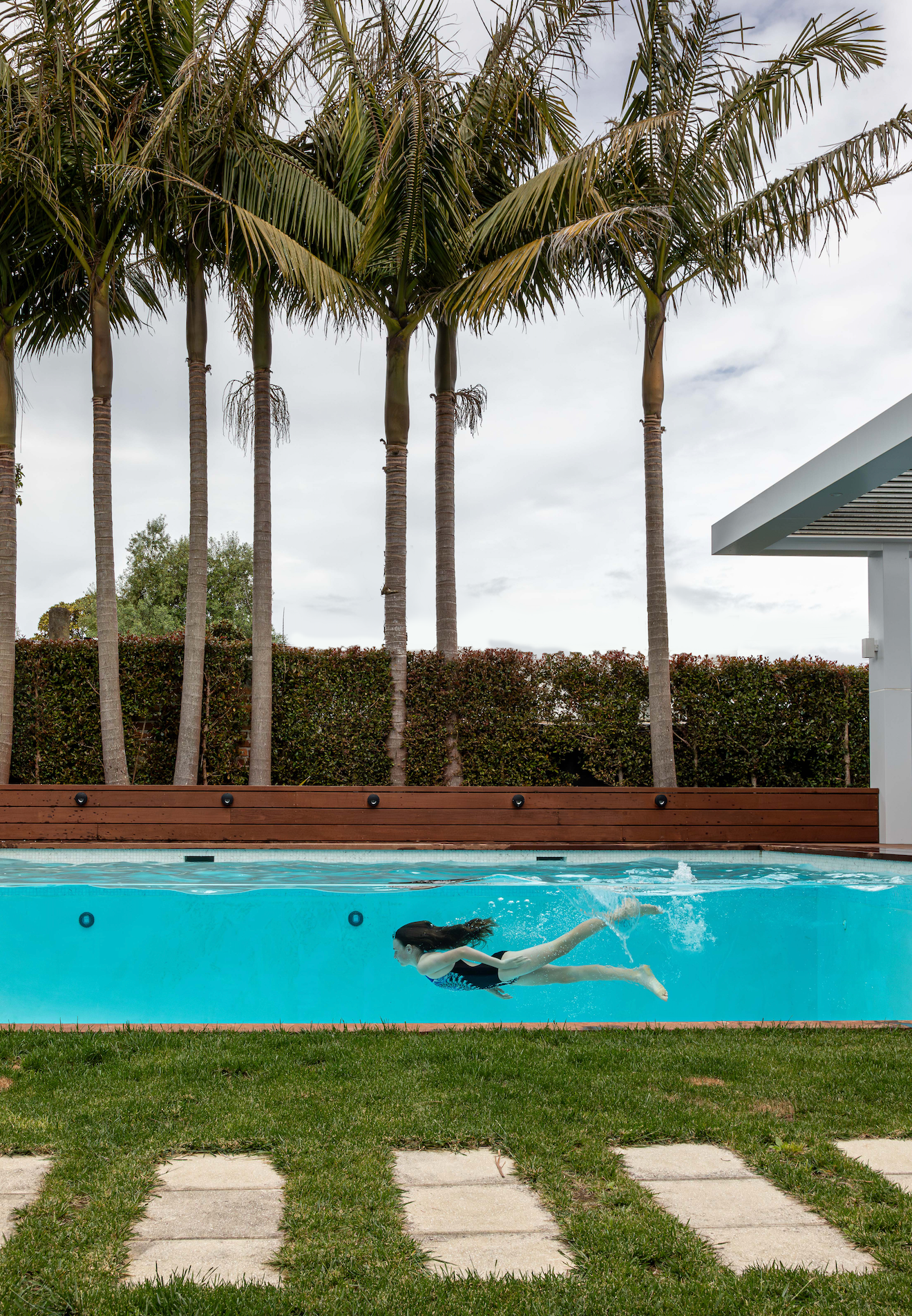 A woman in a black swimsuit floats on her back in a small, rectangular above-ground pool surrounded by lush green tropical plants. The pool has clear sides and a wooden deck on one side with a blue-striped towel draped over the edge. The setting is serene and secluded.