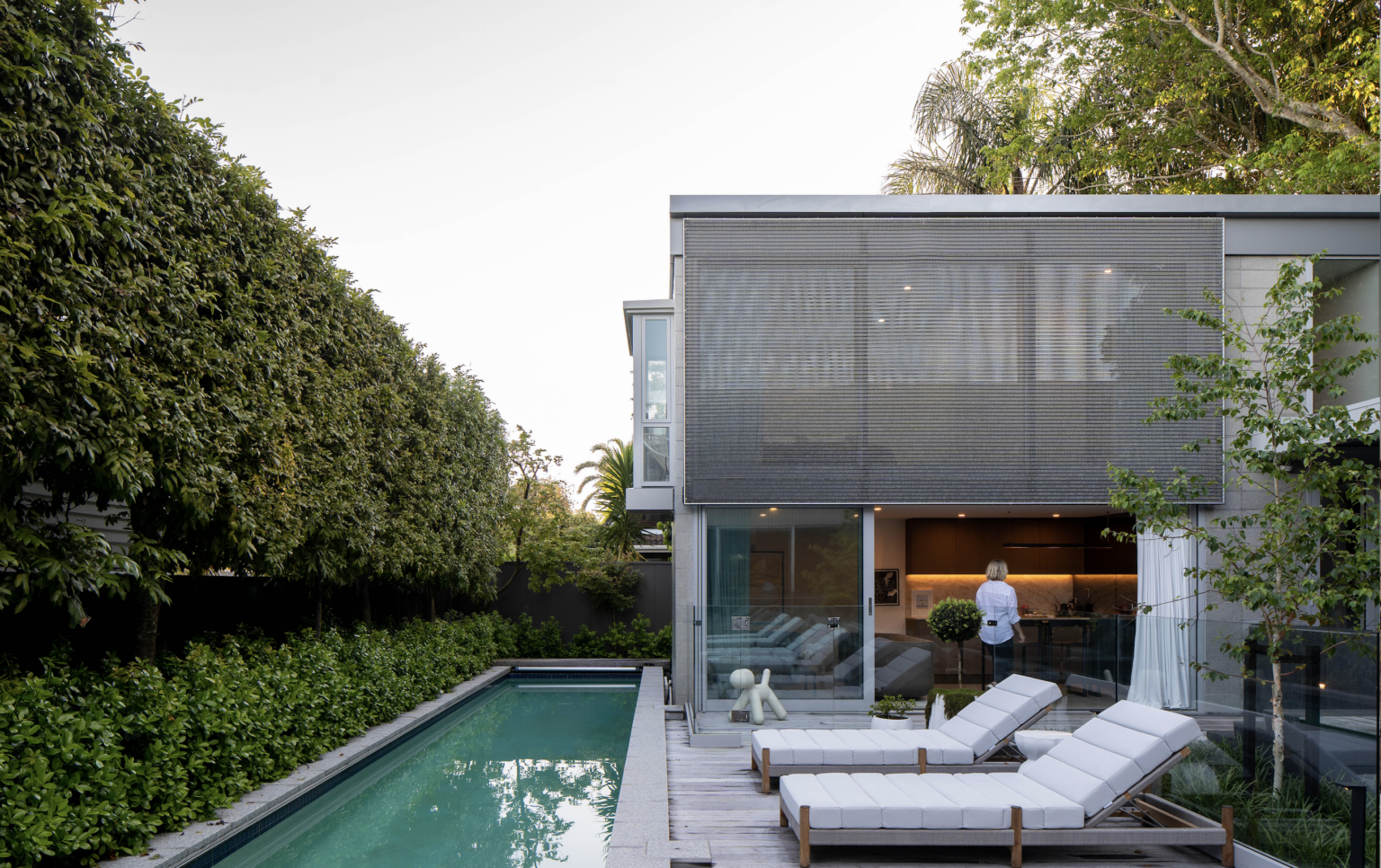 A modern backyard featuring a narrow swimming pool lined with lush green hedges on one side. Adjacent to the pool, there are several white lounge chairs on a wooden deck. The pool is next to a contemporary house with large windows and an upper-level mesh screen. A person is visible inside the house, standing at a kitchen counter. The setting is clean, minimalist, and serene.