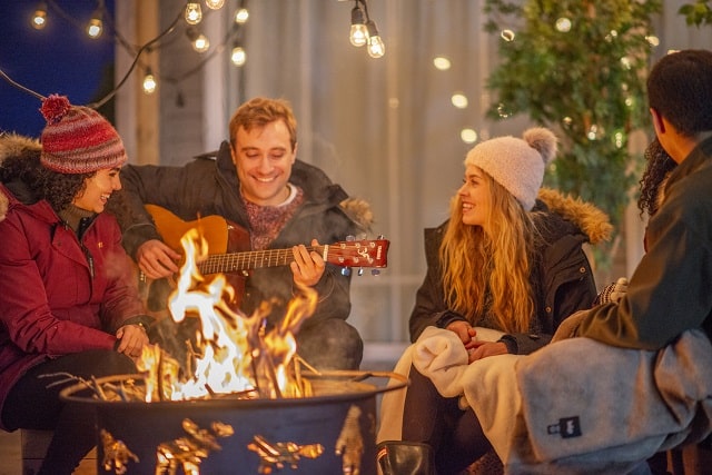 Group of friends wearing thick coats and bobble hats sitting around an outdoor fire while one of them plays guitar.