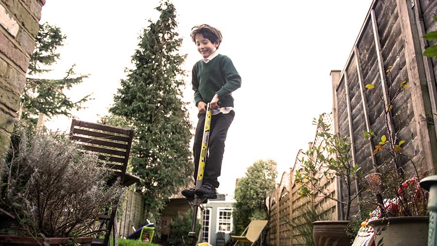  Young boy in school uniform and a helmet bouncing on a pogo stick in the back garden of his house.