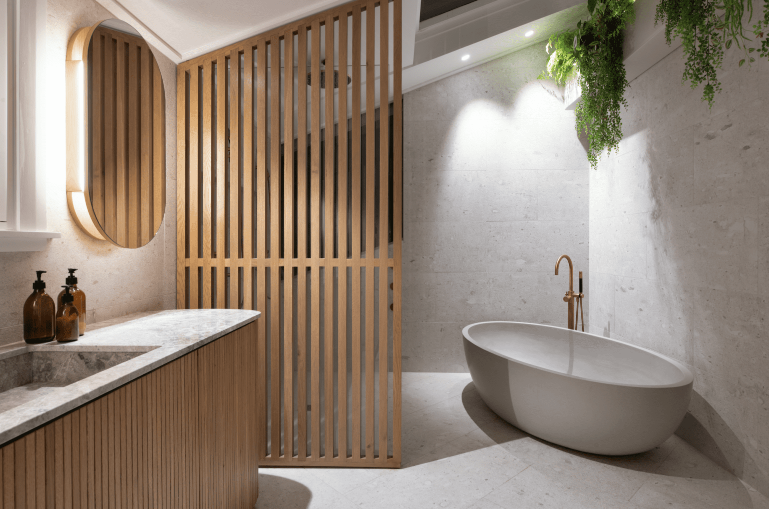 Minimalist bathroom with a freestanding oval bathtub, light grey stone tile walls, and a wood slat privacy divider. The vanity features a marble countertop and a round backlit mirror, while greenery hangs above, adding a touch of nature.