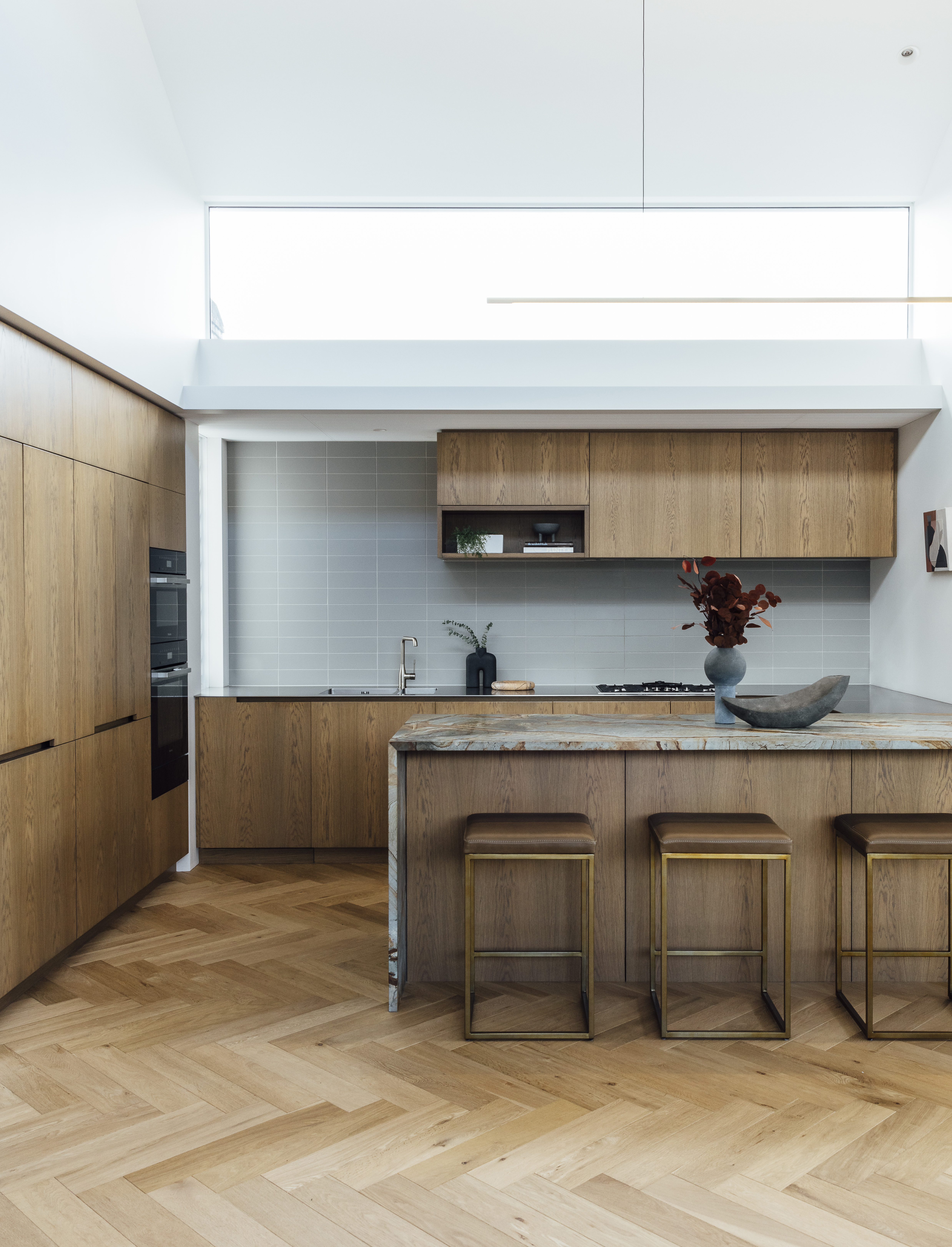 The kitchen offsets the visual complexity of the island’s mare giallo quartzite from Artedomus, the oak veneer cabinetry, and the parquet flooring from Timber Floor Solutions with a simple stainless steel bench, and light grey Antilia tiles from Artedomus. The vases (on the bench and island) and bowl (on the shelf) are by 101 Copenhagen from Frobisher Interiors, the Mesa bowl (on the bench) is from Città, the Untitled 30 artwork is by Grace Badar from Melanie Roger Gallery, the Highline pendant is by Rakumba from ECC and the Harriet bar stools are by David Shaw.”