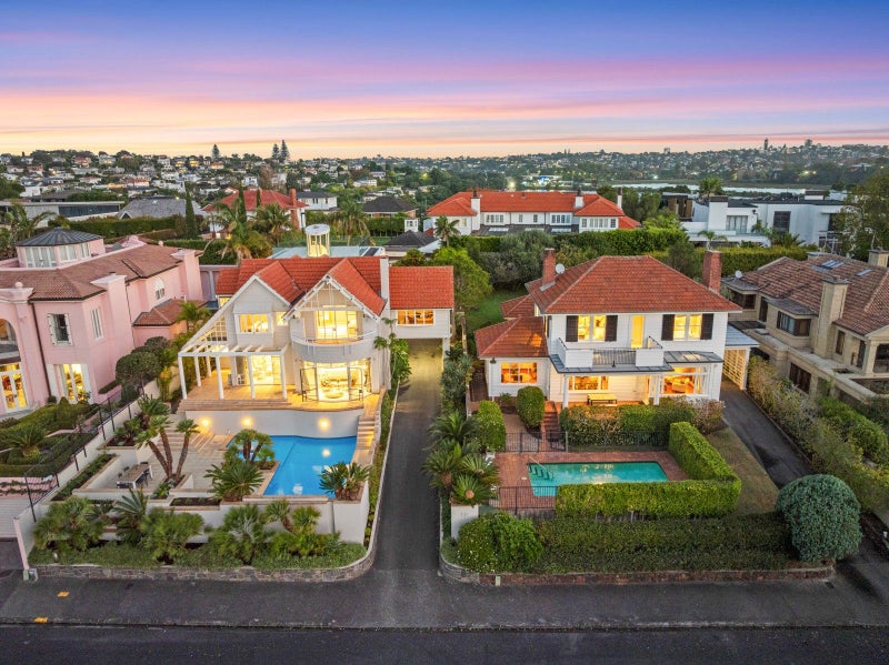 An aerial twilight view of two substantial brick homes with swimming pools, manicured gardens, and red-tiled roofs nestled among tree-lined suburban streets, with the city skyline visible in the distance.