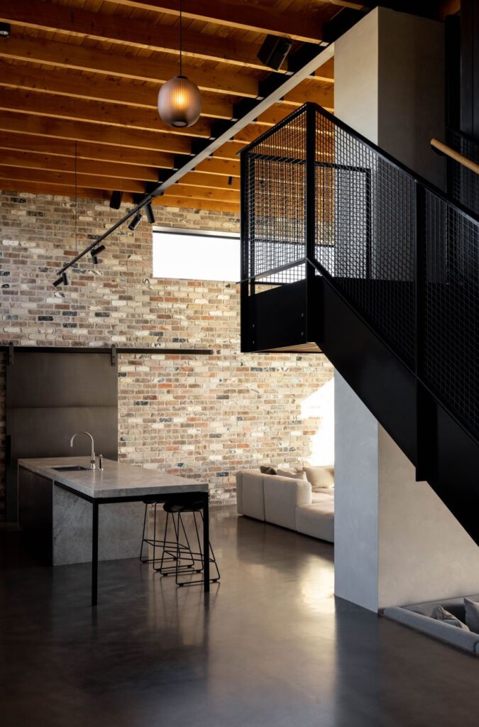 A kitchen and lounge with exposed brick walls and rafters and a black staircase.