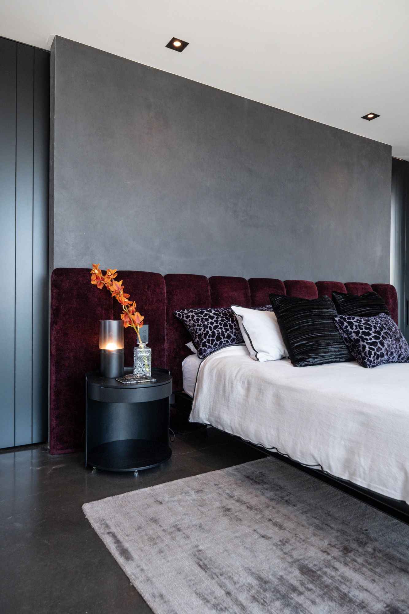 Rich red upholstered headboard adds a rich layer of colour and luxury. An orange flower sits proudly on the black bedside table. Different coloured and textured throw cushions bring the whole room together.