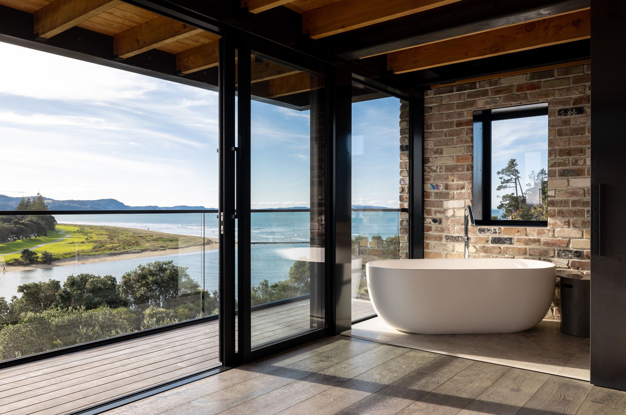 A room with exposed brick walls and rafters with large windows looking out to a sea view.