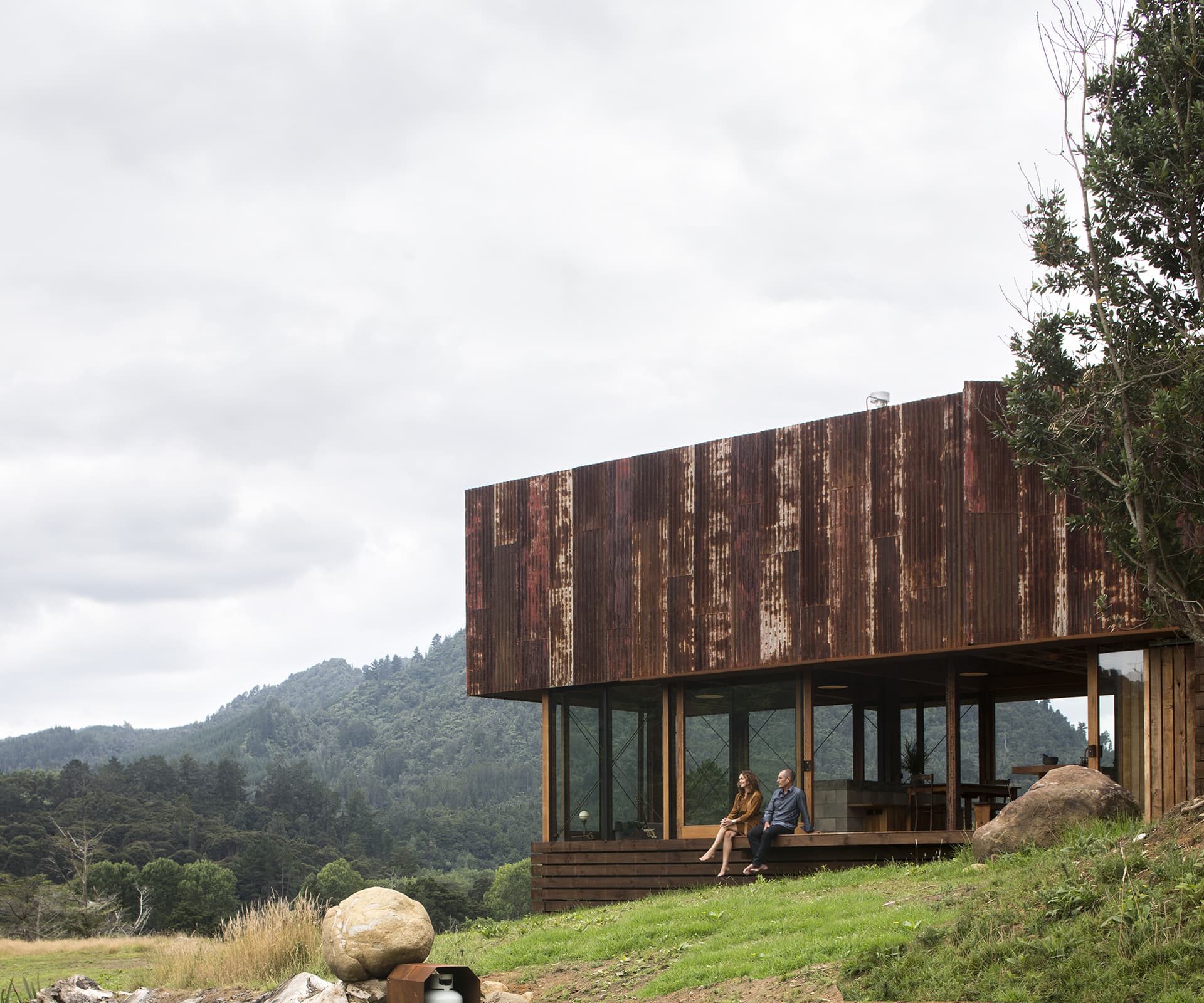 Husband and wife sitting on the deck of their rustic home with a corrugated iron roof and a valley of trees in the background