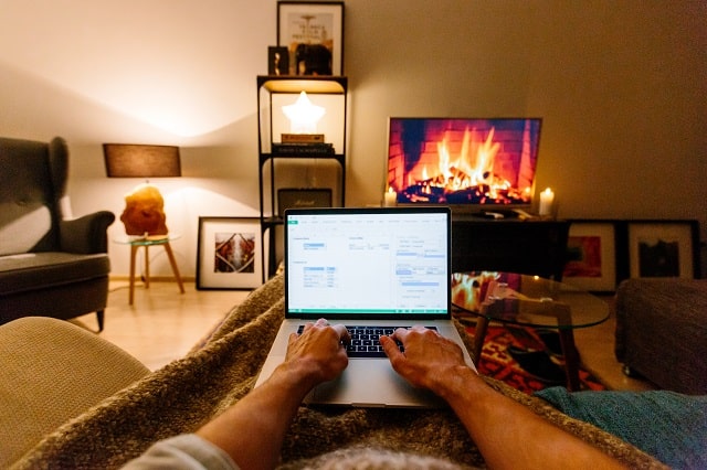 Person working on their laptop, wrapped in a blanket with a TV showing a video of a wood fire.