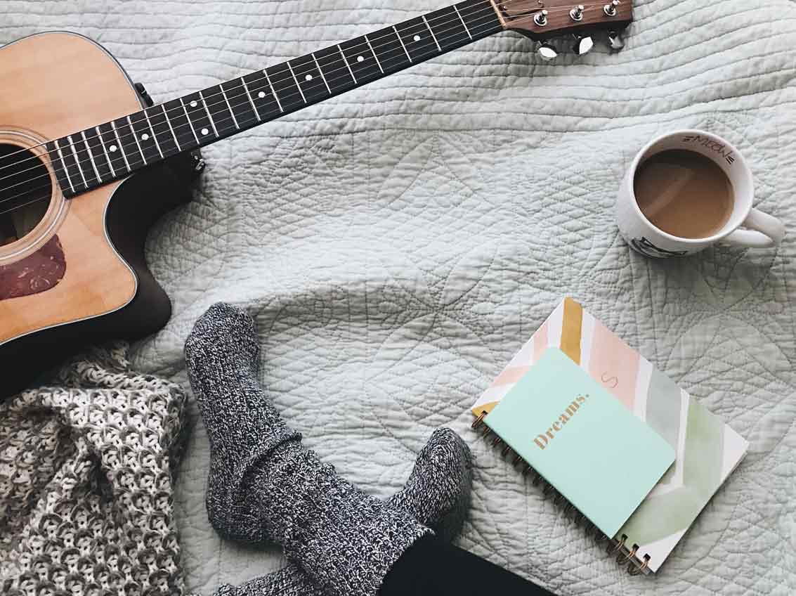 Person wearing woolly socks sitting on a bed with a cup of coffee, some notepads and an acoustic guitar.
