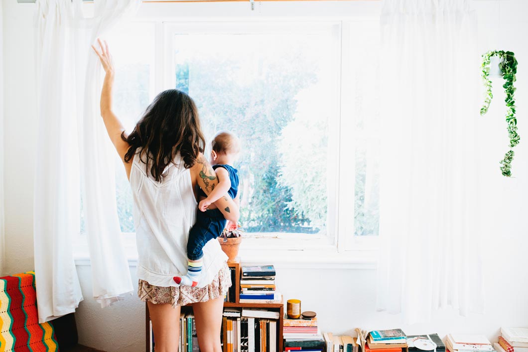 Young mother opening the curtains of her New Zealand home while holding her toddler.