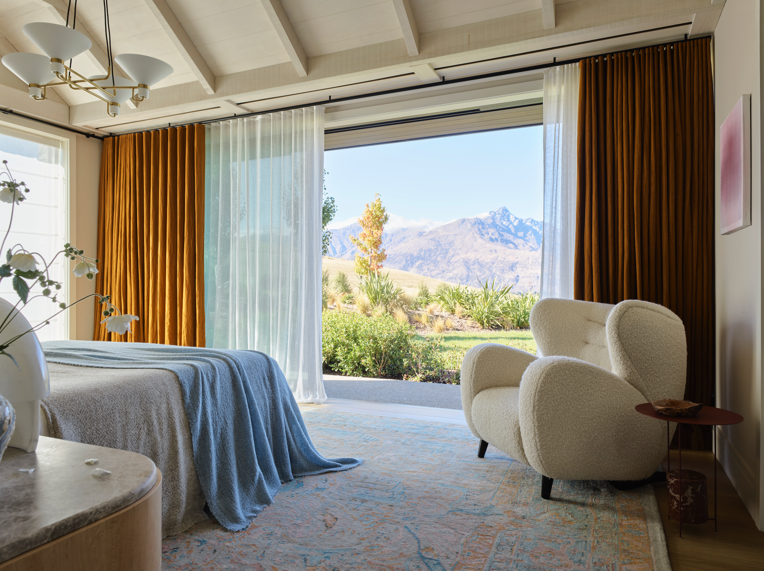 A master bedroom with large windows revealing a stunning mountain view. The room features rich orange curtains, sheer white drapes, a cozy bed with a blue blanket, and a plush white armchair beside a small side table. The décor includes soft, neutral tones and a spacious, airy feel, with natural light flooding the room.