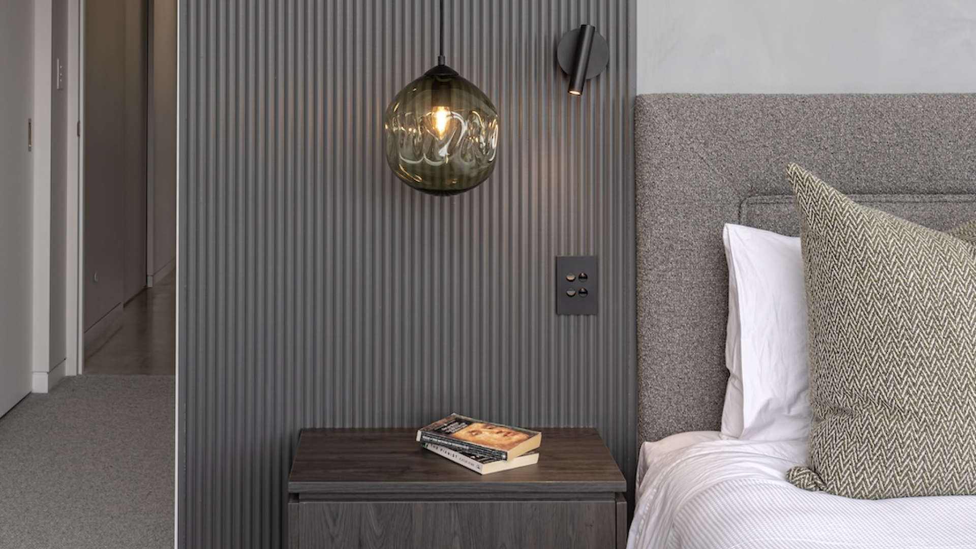 Modern bedroom with a minimalist design, featuring a grey textured accent wall with vertical lines, a hanging glass pendant light, a wall-mounted reading light, and a dark wooden nightstand with a book. The bed has a grey upholstered headboard and white bedding with a patterned throw pillow.