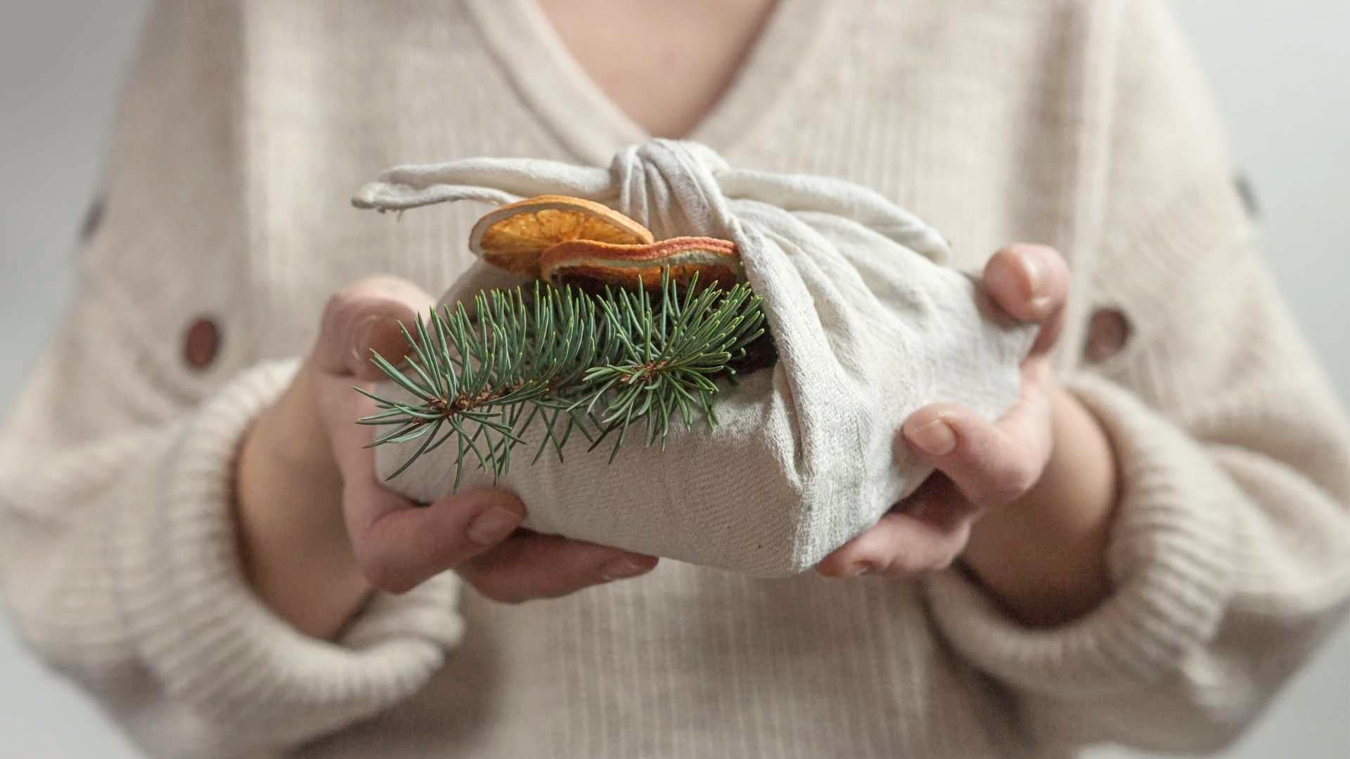 Woman in a beige jumper holding out a gift wrapped in cloth with a sprig of Christmas tree inserted in it. 