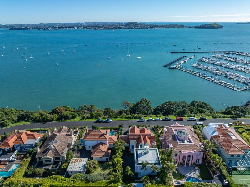 An aerial view of Paritai Drive's clifftop homes with red-tiled roofs lining the waterfront, their large sections separated by manicured hedges, overlooking the bright turquoise water and moored boats below