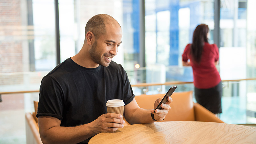 Man sitting at a table with a coffee smiling as he scrolls on his refurbished phone.