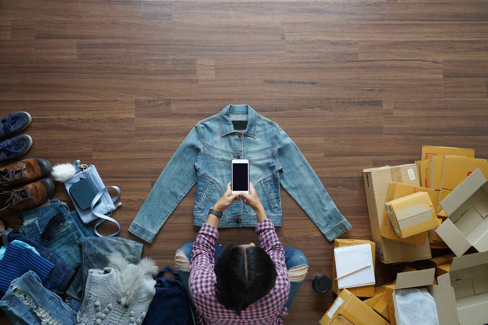 Woman sitting on the floor taking a photo of a denim jacket that she is going to sell on Trade Me. Other items on the floor around her include shoes, bags, a jumper and cardboard boxes she is going to use for shipping.