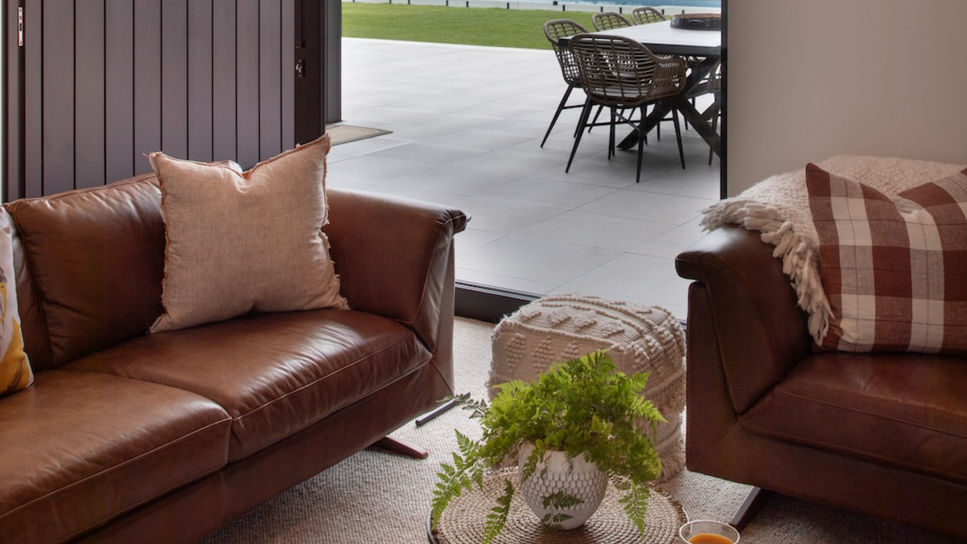 Contemporary living room with leather sofas, neutral cushions, and a round wooden coffee table with a plant and candle. Large sliding glass doors open to a spacious patio with outdoor seating, overlooking a green lawn and distant trees.