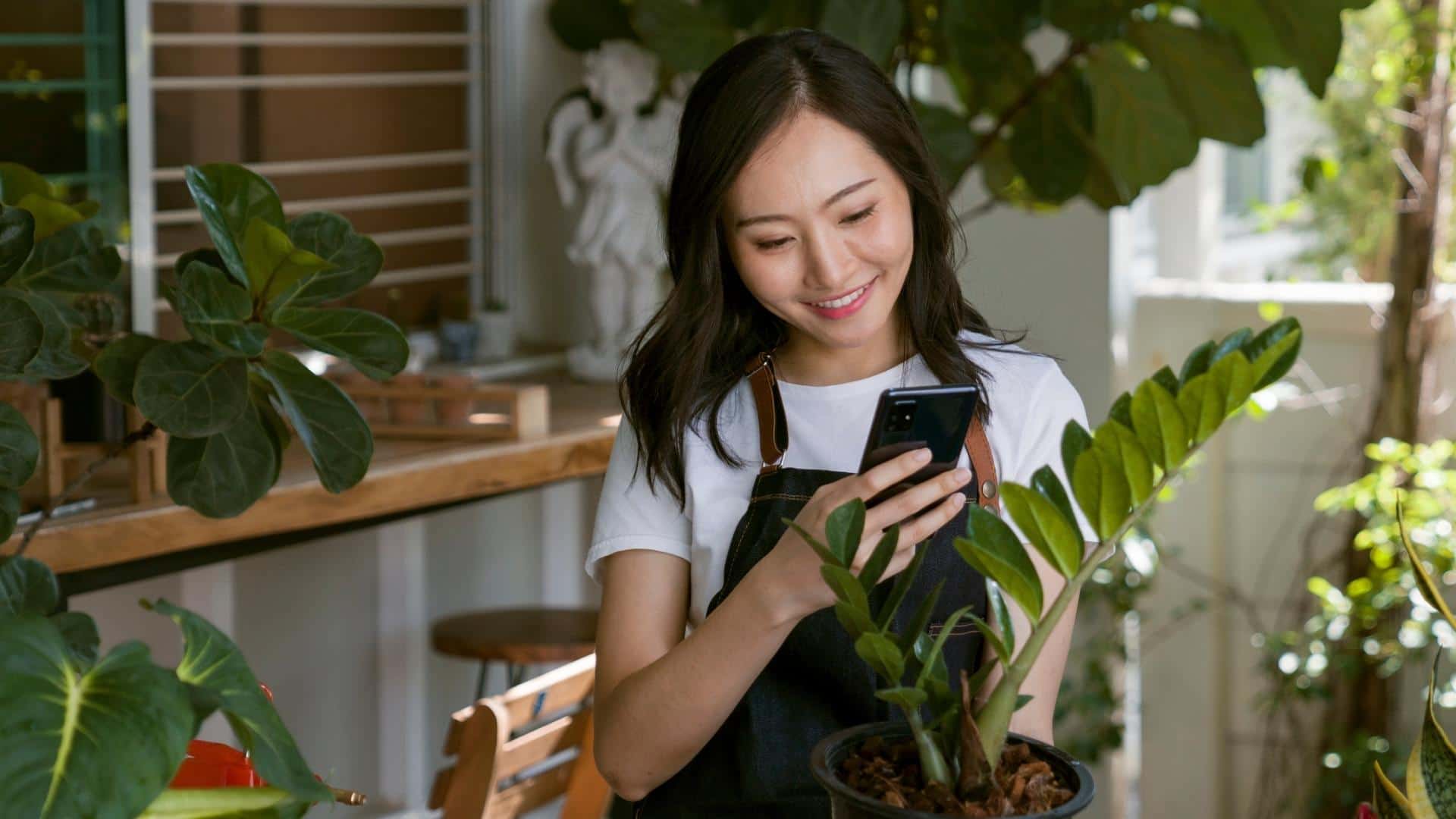 A woman dressed in a white shirt and overalls is surrounded by potted plants and she takes a photo of one in front of her with her phone.