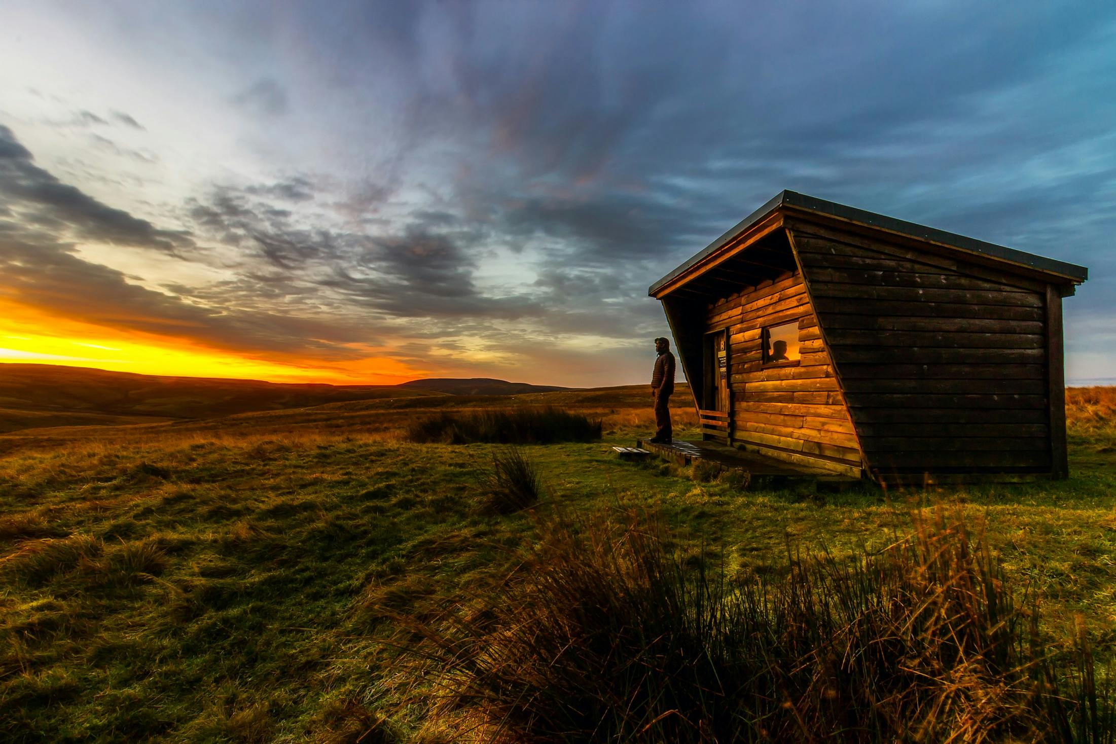 Outside of small log cabin and a person standing in front watching the sun set