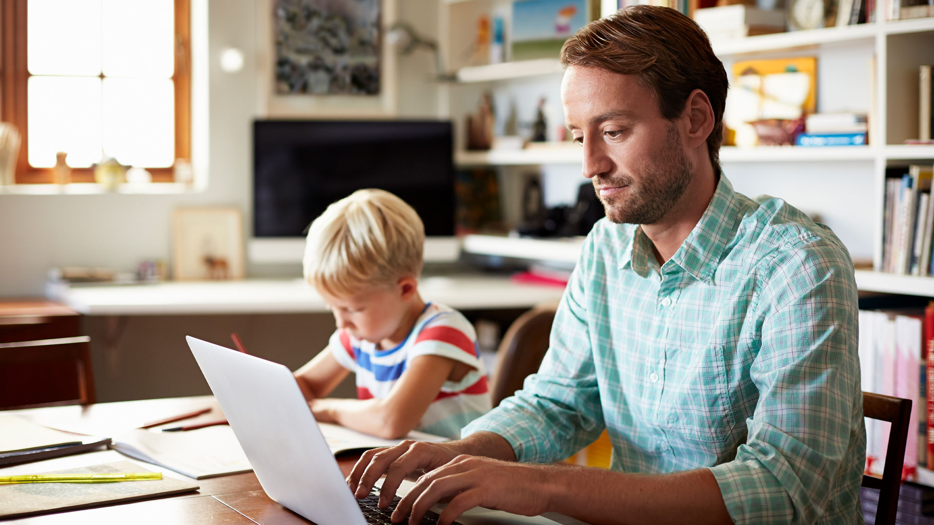 Landlord creating tenancy paperwork on a laptop at home while son does homework in the background.