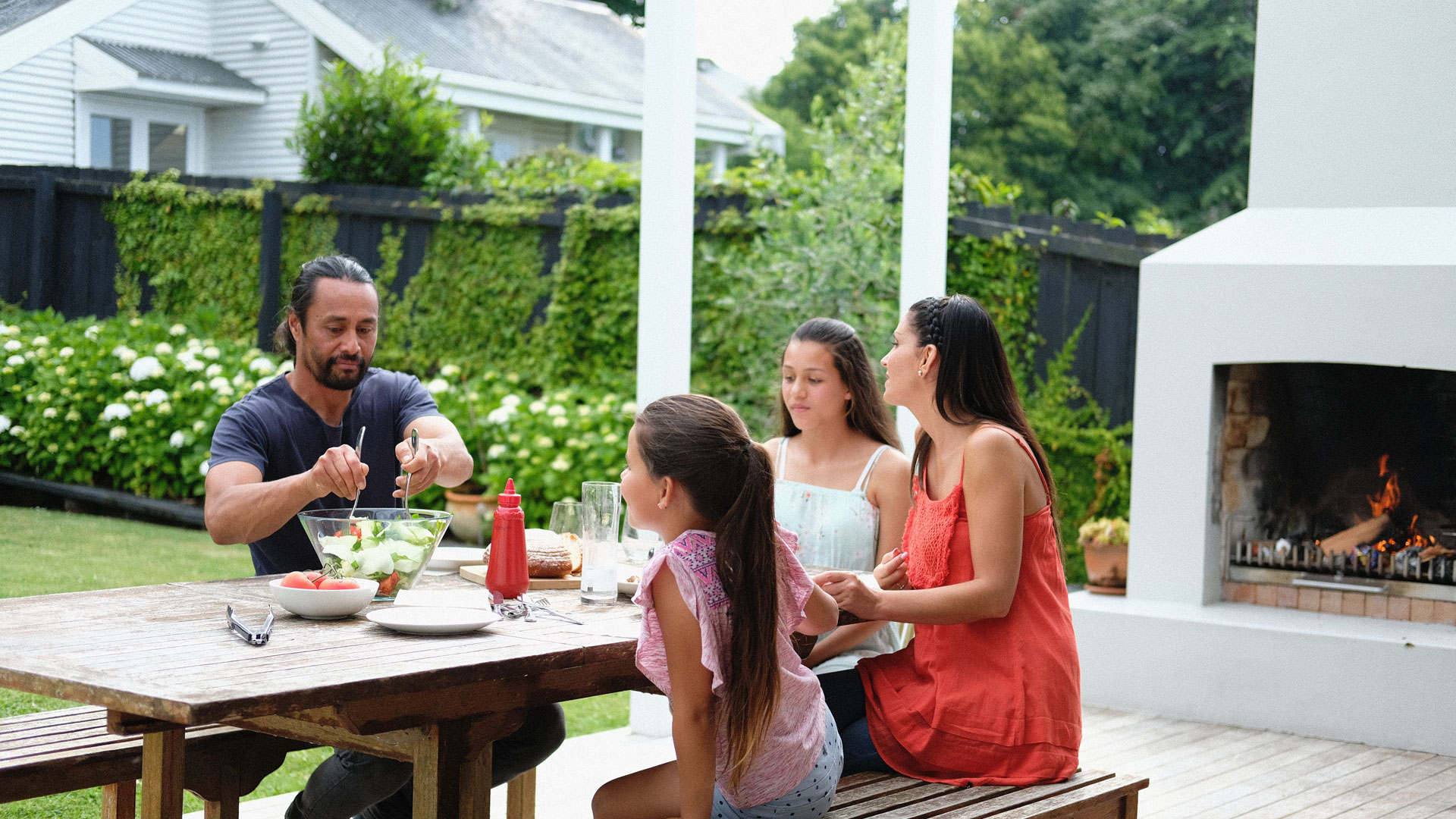 A family in NZ having a meal outdoors in front of a fire in their garden.