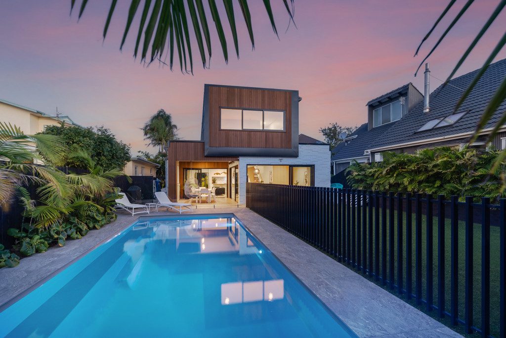 The image showcases a tranquil garden surrounding the modern two-story house during twilight. The lush greenery, vibrant flowers, and well-maintained landscaping create an inviting outdoor space. A swimming pool in the foreground reflects the sky’s colors, adding to the serene ambiance. Overall, it suggests a harmonious blend of nature and architecture.