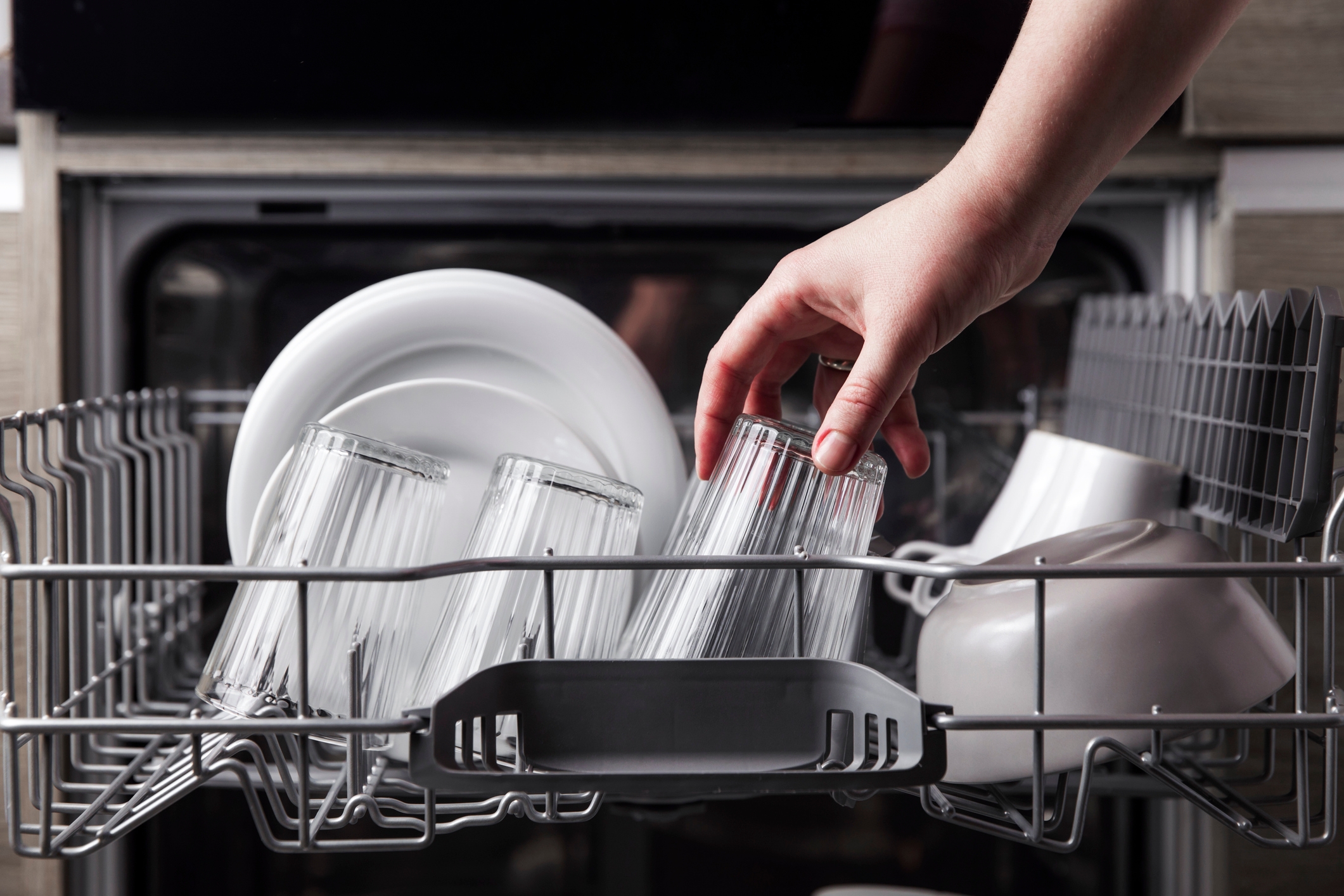 A hand reaching into the top drawer of a dishwasher, picking up a tumbler glass.