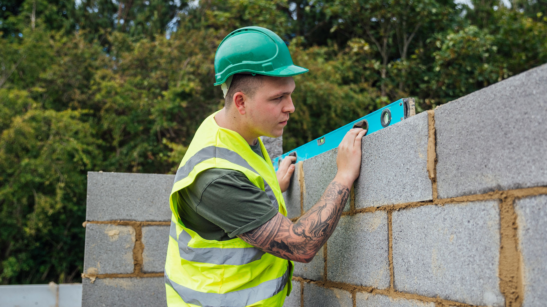 Young man in high vis and a hard hat laying bricks on a construction site.