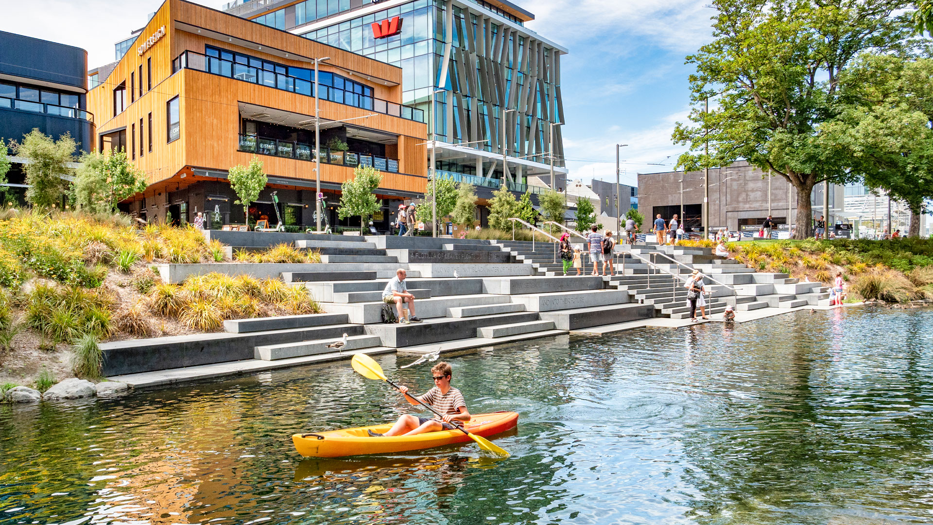 A photo of the Avon river in central Chrischurch. A young boy paddles a kayak down the river, while other people sit on the bank.