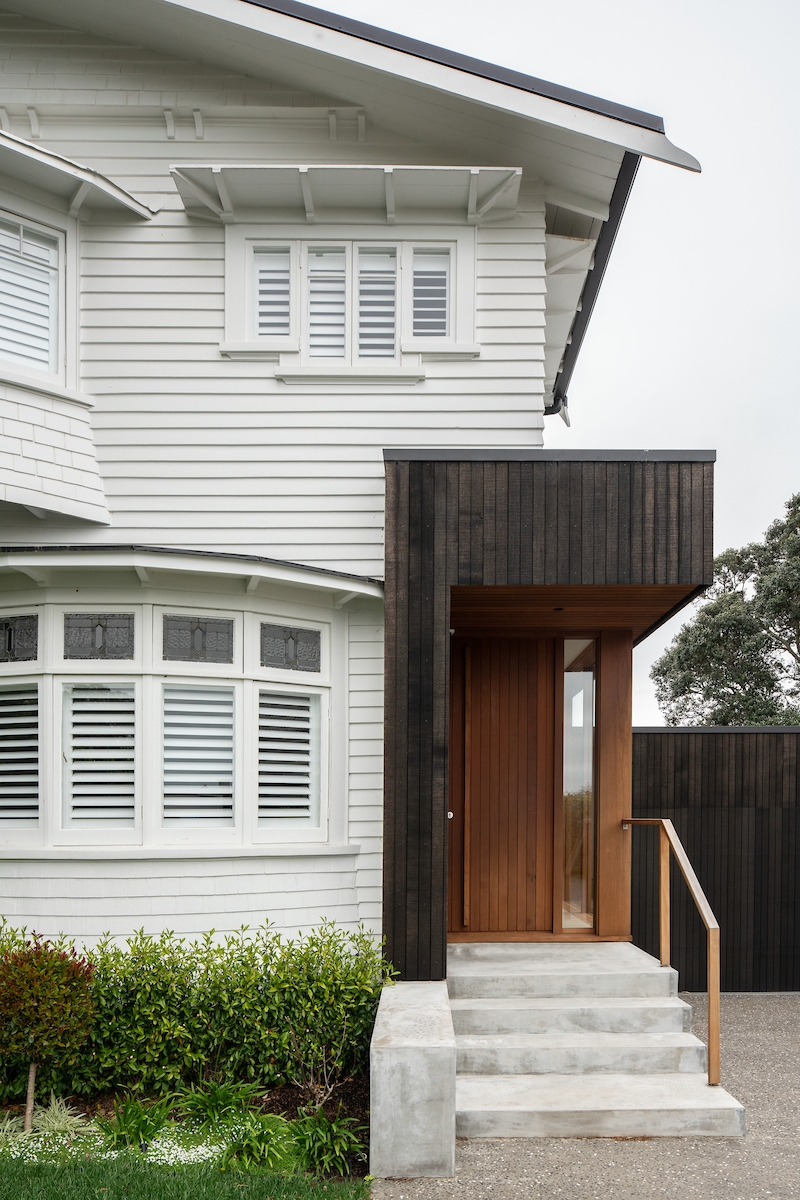 A modern house entrance featuring a combination of white siding and dark wood paneling. The door is made of light wood with a glass panel, set within a dark wood frame that extends above the door to form an overhang. There are three concrete steps leading up to the door, flanked by a metal railing on one side. The house has traditional white-framed windows, and there’s a visible downspout on the left side of the image. A neatly trimmed lawn is in the foreground.