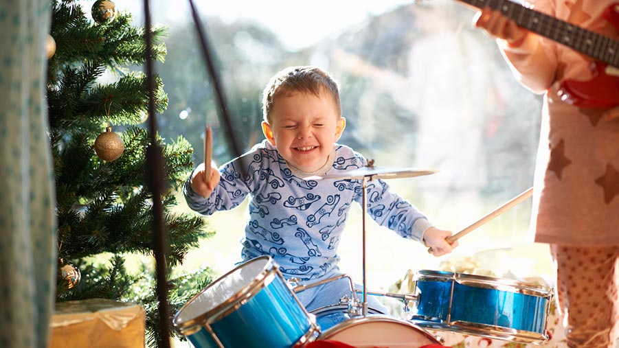 Young boy smiling while playing a child size drum kit as their parent plays guitar in the corner.
