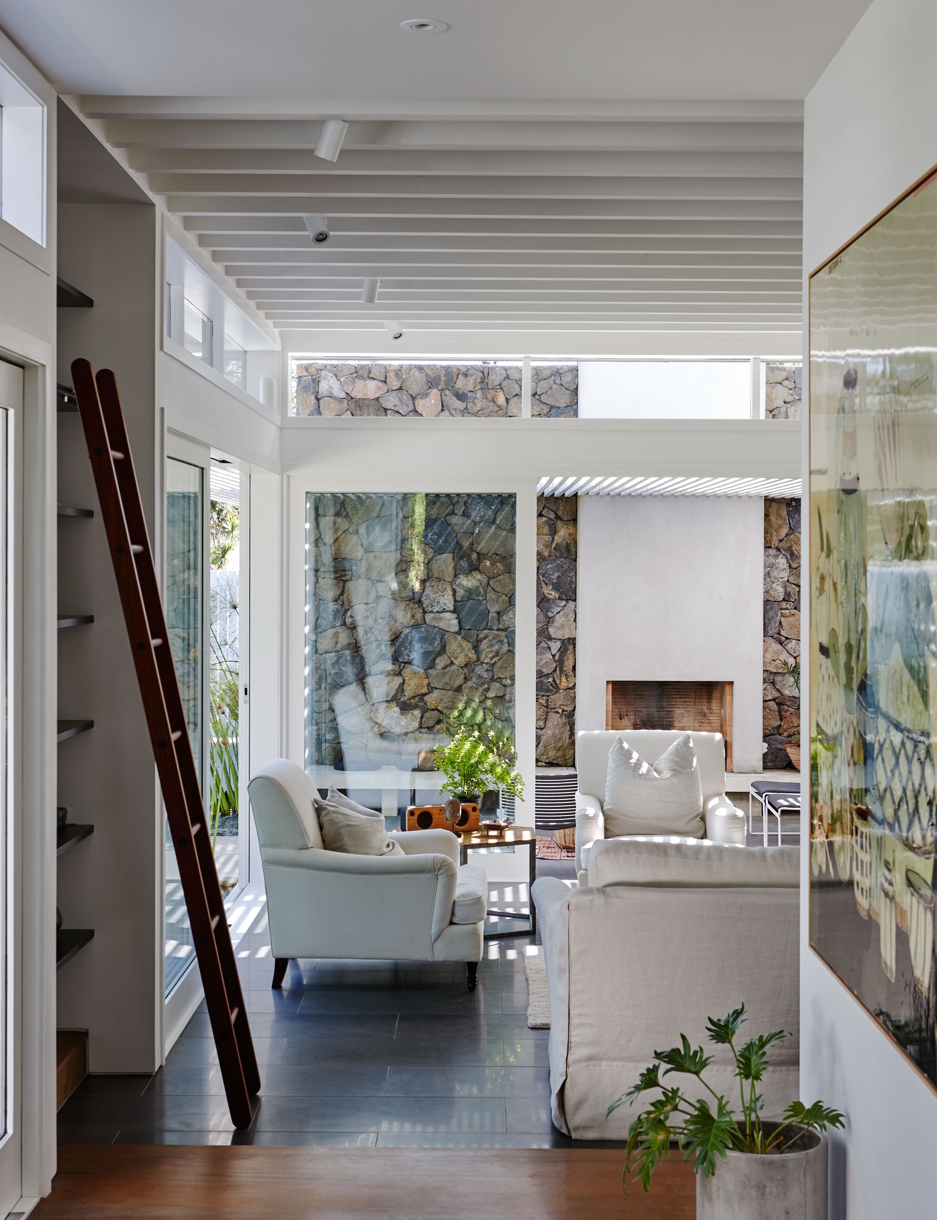 The image depicts an interior space in a modern house. A hallway with white walls and a dark hardwood floor leads to a living area. In the living area, there are two armchairs—one white and one grey—along with a small plant. A stone fireplace is visible in the background, and large windows provide natural light and views of greenery outside. The ceiling features recessed lighting and exposed beams. This image could be relevant for discussions on interior design and home decor.